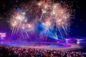 A large crowd gathered at night watching a vibrant fireworks and light display over the water.