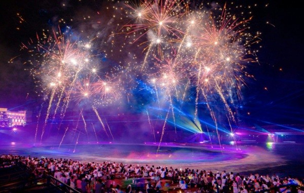 A large crowd gathered at night watching a vibrant fireworks and light display over the water.